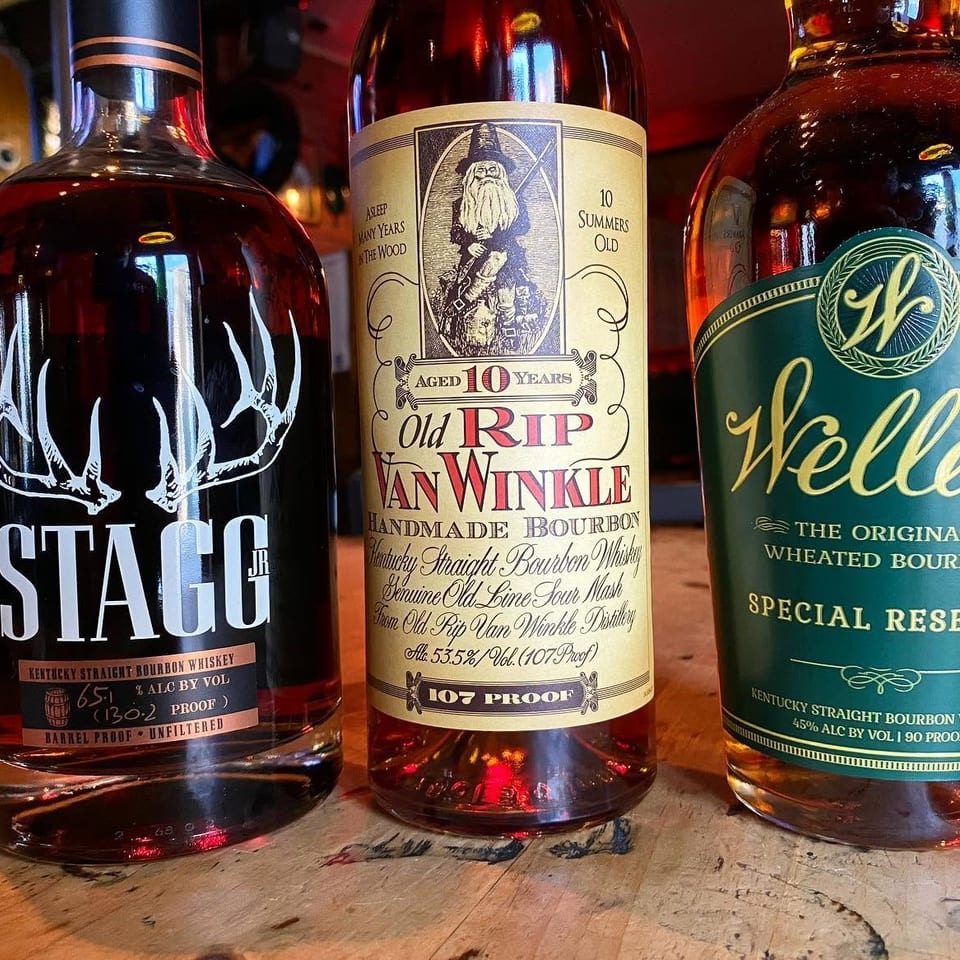 Three bourbon whiskey bottles—Stagg Jr., Old Rip Van Winkle, and Weller—displayed on a wooden bar.